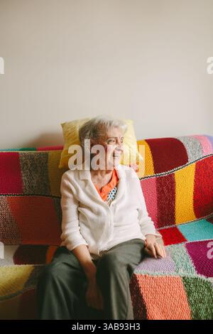 Eine ältere Frau sitzt auf einem bunten, strukturierten Sofa und lächelt warm. Sie verkörpert Wärme und Freude und verleiht der ruhigen Umgebung Charme. Stockfoto