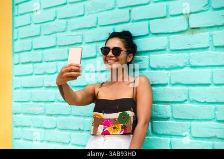 Eine junge Lateinfrau lächelt breit, während sie ein Selfie gegen eine türkisfarbene Ziegelwand macht. Sie trägt Sonnenbrille und legere Kleidung. Stockfoto