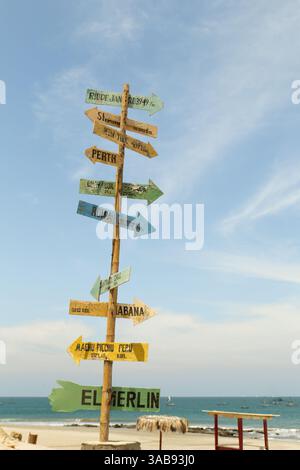 Am Mancora Beach, Peru, befindet sich ein lebendiger hölzerner Wegweiser, der zu Reisezielen auf der ganzen Welt führt. Der klare blaue Himmel und die Meereswellen im Hinterland Stockfoto
