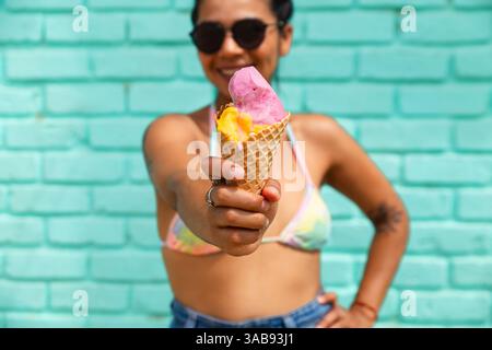 Eine junge Lateinfrau hält in Mancora, Peru, fröhlich eine Eiskugel vor türkisfarbenem Hintergrund. Die Szene fängt ein lebhaftes, sonniges Strandatmosp ein Stockfoto