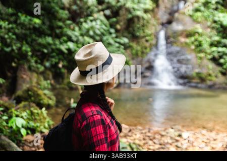 Seitenansicht einer nicht erkennbaren Frau mit Hut und Blick auf einen malerischen Wasserfall im Choco Andino Regenwald in Ecuador. Sie ist in einem Karo gekleidet Stockfoto