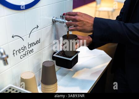 Ein indischer Mann, der frisch gebrühten Kaffee aus einem an der Wand montierten Spender in einem Hotelcafé bei einer KI-Konferenz in Amsterdam in eine Papiertasse gießt. Stockfoto