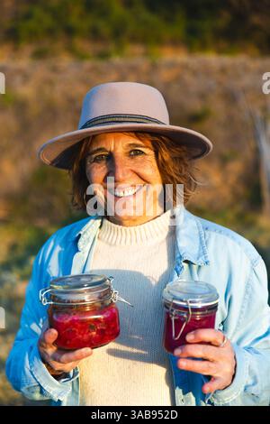 Eine fröhliche Frau mit Hut und Jeansjacke, die in Barcelona, Spanien, Gläser mit selbstgemachten Konserven hält. Das Sonnenlicht verstärkt ihren fröhlichen Ausdruck Stockfoto