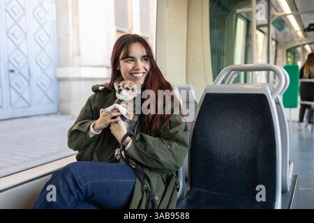 Eine fröhliche Frau sitzt auf einem Straßenbahnsitz und hält ihren kleinen chihuahua, warm in einen Mantel gewickelt. Die moderne Innenausstattung der Straßenbahn schafft eine gemütliche Umgebung für ihre Gäste Stockfoto