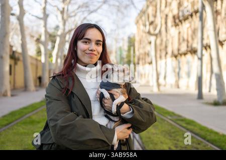 Eine Frau mit langen dunklen Haaren lächelt, während sie ihren chihuahua hält, und genießt einen sonnigen Tag in einer von Bäumen gesäumten Straße in einem gemütlichen weißen Pullover und grünem jac Stockfoto
