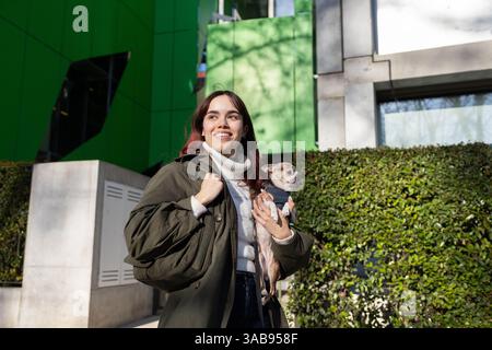 Eine Frau in einer grünen Jacke hält einen kleinen chihuahua und genießt einen sonnigen Tag vor einem modernen Gebäude mit grünen Paneelen und üppigem Grün im Rücken Stockfoto