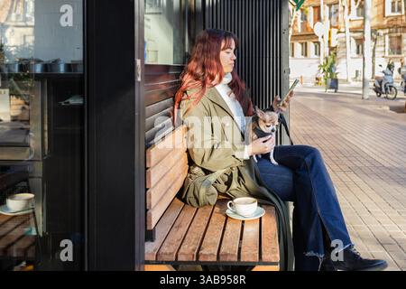 Eine Frau sitzt auf einer Bank vor einem Café und schaut auf ihr Handy, mit einem kleinen chihuahua auf dem Schoß. Sie trägt einen grünen Mantel und Jeans, während sie eine Kaffeetasse trägt Stockfoto