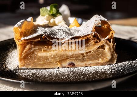 Ein Stück traditioneller österreichischer Apfelstrudel, mit Puderzucker bestäubt und mit Schlagsahne und Minze begleitet, auf einer dunklen Keramikplatte unter nat Stockfoto