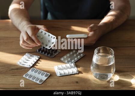 Ein Mann, der eine Blisterpackung mit Pillen hält, während er ein Smartphone benutzt, mit mehreren Päckchen und einem Glas Wasser auf einem Holztisch, was den Drogenmann hervorhebt Stockfoto