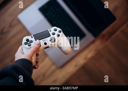 Nahaufnahme einer Person, die einen Gaming-Controller über einem Laptop auf einem Holztisch hält. Die Szene zeigt die Integration von Gaming und Technologie, Highlig Stockfoto