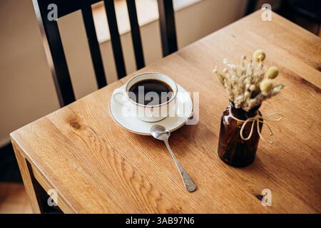 Eine gemütliche Szene mit einer Kaffeetasse auf einem Holztisch am Fenster in einem Café. Eine kleine Vase mit getrockneten Blumen verleiht einen Hauch von rustikalem Charme, perfekt für Stockfoto