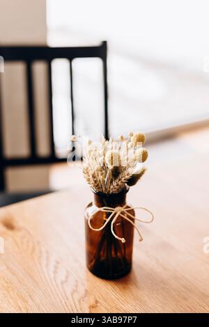 Eine Glasvase, die mit Garn verbunden ist, hält einen Strauß getrockneter Blumen auf einem Holztisch in einem Café. Weiches Licht verstärkt den rustikalen Charme und schafft eine gemütliche Atmosphäre Stockfoto
