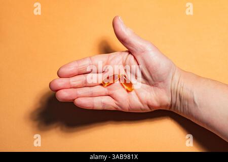 Eine Nahaufnahme einer offenen, nicht erkennbaren Hand, die zwei durchscheinende Omega-3-Kapseln vor einem leuchtenden orangefarbenen Hintergrund hält. Das Bild emphasi Stockfoto