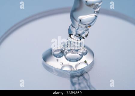 Nahaufnahme der feinen Bewegung einer klaren, viskosen Flüssigkeit mit goldenen Flocken, die von einer Kosmetikpipette auf einen Reflektor fallen Stockfoto