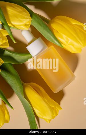 Eine flache Komposition mit einer mattierten Tropfenkosmetik aus Gesichtsserum, umgeben von leuchtend gelben Tulpen auf einem warmen, neutralen Hintergrund. Perfekt Stockfoto