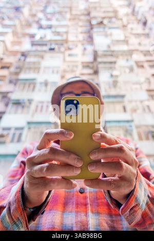 Von unten aus benutzt ein nicht erkennbarer Mann mit Hut ein Smartphone vor einer dichten Kulisse in Hongkong. Stockfoto