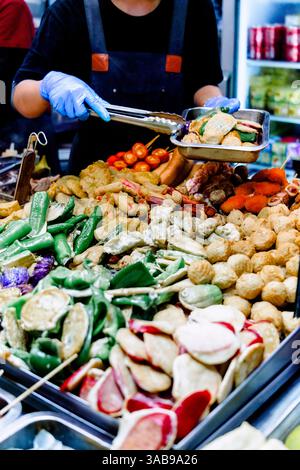 Ein nicht erkennbarer Straßenverkäufer in Hongkong, der geschickt eine Auswahl an bunten frittierten Gemüsesorten und Tofu aus einem geschäftigen Essen serviert Stockfoto