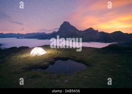 Ein leuchtendes Zelt befindet sich auf einem grasbewachsenen Hügel unter einem Sternenhimmel, umgeben von nebelbedeckten Bergen in den Pyrenäen. Die heitere Szene fängt das wesentliche ein Stockfoto