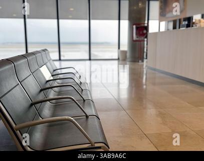Leerer Sitzbereich am Flughafen mit Metallbänken, mit Blick auf den Asphalt durch große Fenster. Der Boden ist poliert und spiegelt den ruhigen, offenen Raum wider Stockfoto