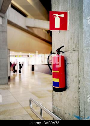 Ein roter Feuerlöscher, der an einer Betonwand in einem Flughafenterminal montiert ist. Ein Sicherheitsschild darüber sorgt für eine gute Sicht. Verschwommener Hintergrund mit Personen W Stockfoto