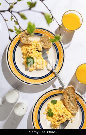 Zwei Teller mit cremigem Rührei mit Toast, garniert mit Kräutern, serviert zusammen mit Gläsern frischen Orangensafts. Ein perfekter, heller Morgen. Stockfoto