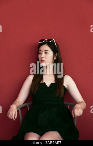 Eine junge asiatin in schwarzem Kleid und Sonnenbrille sitzt vor einer leuchtend roten Wand. Ihr ruhiges Auftreten und die kontrastfarbenen Farben sorgen für ein markantes VI Stockfoto