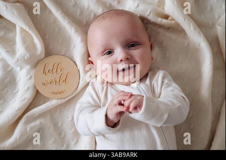Ein lächelnder Junge liegt auf einer weichen, cremefarbenen Decke. Neben ihm trägt eine hölzerne Tafel die Aufschrift Hello World, auf der die Wärme und Unschuld der frühen Kindheit festgehalten werden Stockfoto
