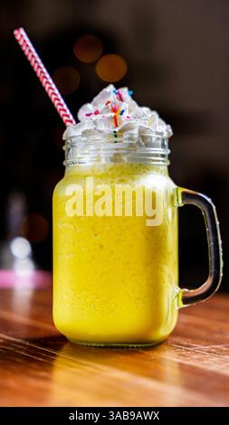 Ein lebendiger Mango-Smoothie mit cremiger Schlagsahne und bunten Streuseln, serviert in einem mauerglas mit gestreiftem Stroh auf einem hölzernen Kaffeetisch. P Stockfoto