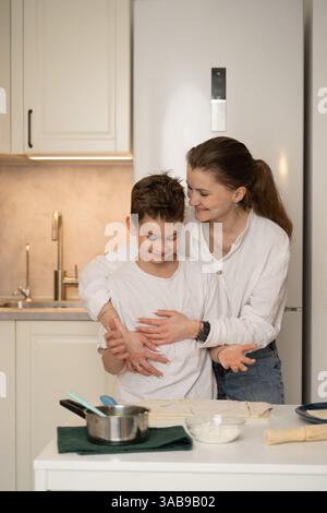Eine Mutter umarmt ihren Sohn liebevoll, während sie in ihrer gemütlichen Küche eine Mahlzeit zubereitet. Die Szene fängt einen warmen Familienmoment mit Backutensilien auf dem C ein Stockfoto