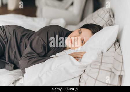 Eine junge Frau in bequemer Loungewear liegt auf einem weißen Bett und legt ihren Kopf auf weichen Kissen, während sie sanft lächelt. Die warme und gemütliche Atmosphäre ist hoch Stockfoto