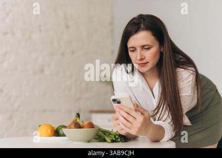 Ernährungswissenschaftler untersucht das Smartphone, während er sich mit frischem Obst und Gemüse auf den Tisch lehnt. Konzept der gesunden Ernährung, ausgewogene Ernährungsberatung und Lebensführung Stockfoto