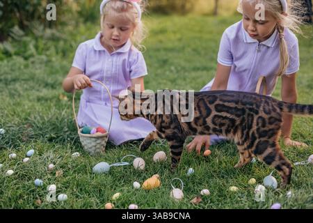 Zwei Kinder in lila Outfits genießen eine Ostereierjagd in einem Garten, begleitet von einer neugierigen Katze. Farbenfrohe Eier umgeben sie auf dem üppig grünen Gras Stockfoto