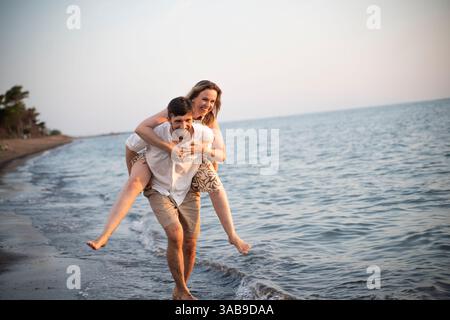 Ein fröhliches Paar genießt einen verspielten Tag an einem italienischen Strand, wobei ein Partner dem anderen einen Huckepack-Ritt gibt. Das ruhige Meer und der weiche Sand bilden ein pe Stockfoto
