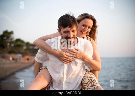 Ein fröhliches Paar genießt einen verspielten Tag an einem italienischen Strand, wobei ein Partner dem anderen einen Huckepack-Ritt gibt. Das ruhige Meer und der weiche Sand bilden ein pe Stockfoto