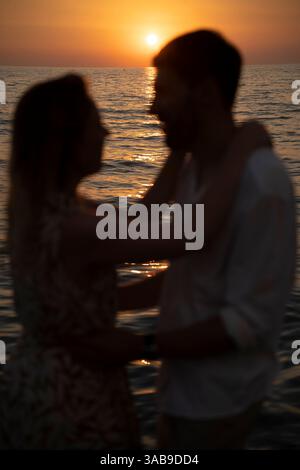 Ein Paar umarmt einen italienischen Strand während Sonnenuntergang. Ihre Silhouetten harmonieren mit den sanften Wellen und dem lebendigen Himmel. Ein ruhiger, intimer Moment bei den s Stockfoto