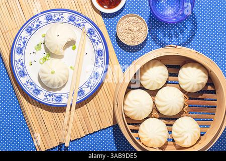 Von oben werden zwei bao-Brötchen auf einem eleganten blauen Teller mit Essstäbchen serviert, begleitet von einem Bambusdampfer mit frisch zubereiteten Brötchen. Die Stockfoto