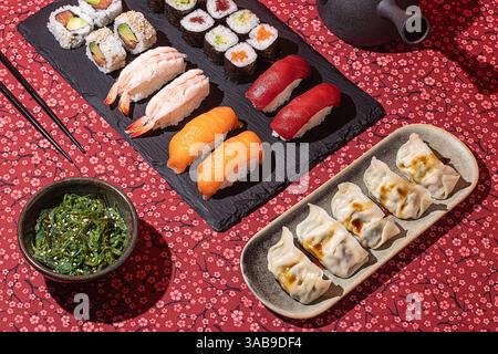 Flacher Lay eines traditionellen japanischen Essens mit Nigiri, Maki-Rollen und Gyoza, präsentiert auf einer roten Blumendecke mit dramatischen Schatten Stockfoto