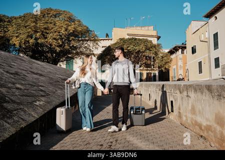 Ein junges Paar geht Hand in Hand mit Koffern und erkundet die charmanten Straßen Mallorcas, Spanien, während sie nach ihrem idealen Ferienhaus suchen. Sonntags Stockfoto