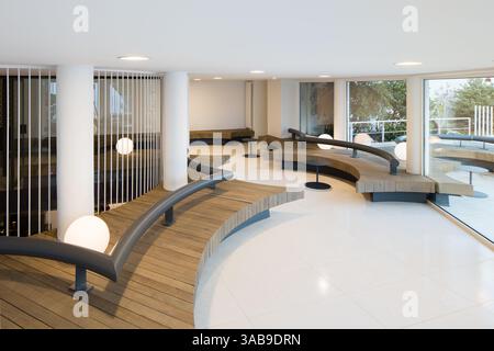 Gebogene Holzbänke mit Metallakzenten in einem Club-Innenbereich. Große Fenster und runde Lichter bieten ein modernes Ambiente, ideal für Bar oder Club. Stockfoto