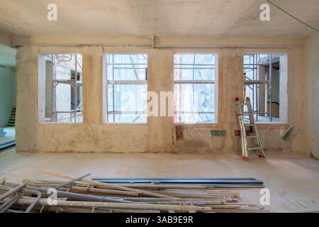 Das Innere eines im Bau befindlichen Gebäudes mit großen Fenstern, Gerüsten und einer Metalltreppe. Der Raum ist mit Baumaterialien gefüllt. Stockfoto