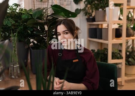 Eine junge Floristin mit schwarzer Schürze, umgeben von verschiedenen Pflanzen. Sie sitzt an einem Holztisch mit Regalen, die mit Pflanztöpfen gefüllt sind Stockfoto