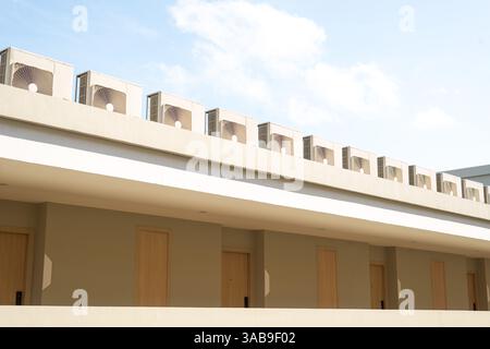 Eine Reihe von Klimaanlagen auf dem Dach eines modernen Gebäudes vor einem klaren blauen Himmel in Thailand. Das beige Äußere mit Holztüren schafft einen Mini Stockfoto