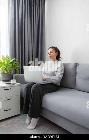 Eine Frau sitzt auf einem bequemen grauen Sofa in einem gut beleuchteten Wohnzimmer und arbeitet an einem Laptop. In der Nähe befindet sich eine Topfpflanze und eine Kaffeetasse auf einem weißen Beistelltisch. Stockfoto
