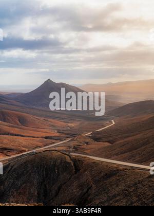 Ein atemberaubender Blick auf eine kurvenreiche Straße, die durch die zerklüfteten Berge von Fuerteventura führt. Das warme Sonnenlicht und die dramatischen Wolken heben die Insel hervor Stockfoto