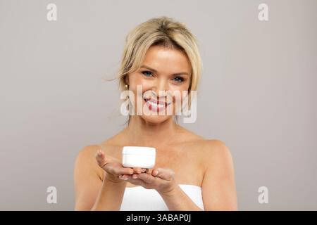 Weiße Frau mittleren Alters lächelt warm, während sie ein weißes Kosmetikglas präsentiert Stockfoto