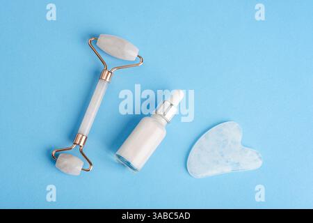 Hautpflegewerkzeuge einschließlich Gesichtsrolle, Serumtropfflasche und Gua-Sha-Stein, angeordnet auf blauem Hintergrund in minimaler, flacher Zusammensetzung für Schönheit Stockfoto