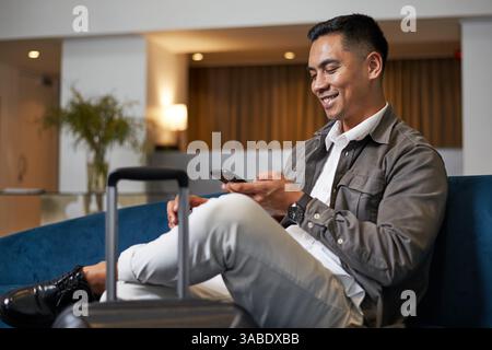 Lächelnder Reisender, der sich während einer Geschäftsreise in der Hotellobby mit dem Smartphone entspannt Stockfoto