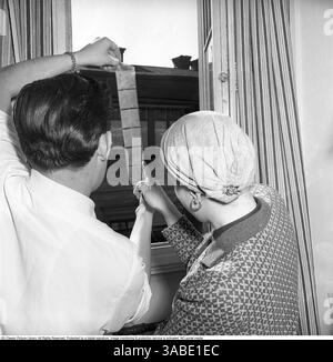Ein Mann und eine Frau, die an einem Fenster stehen und einen Streifen mittelformatiger fotografischer Negativen zum Licht hochhalten, um die Bilder zu untersuchen. Der Mann hält den Streifen mit der rechten Hand, während die Frau mit der linken Hand darauf zeigt, scheinbar die Details bespricht oder überprüft. Dieses Bild gibt einen faszinierenden Einblick in den alten Prozess der Überprüfung von Fotos vor der Entwicklung und zeigt eine Methode, die einst für Fotografen und Enthusiasten unerlässlich war. Es spiegelt eine Ära wider, in der physischer Film sorgfältig behandelt, studiert und geschätzt wurde, bevor er in greifbare Formen verwandelt wurde Stockfoto