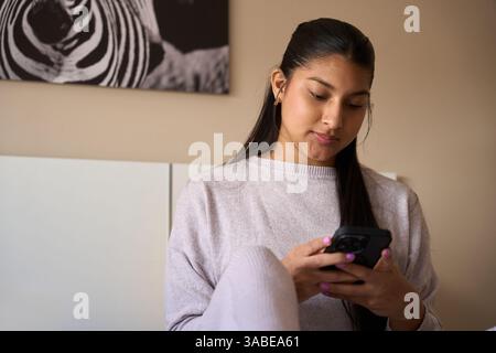 Junge Frau, die ihr Smartphone benutzt und zu Hause auf dem Bett sitzt Stockfoto