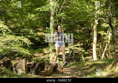 Frau, die auf einem Waldweg läuft, über umfallene Baumstämme geht und einen sonnigen Tag inmitten üppiger grüner Bäume genießt Stockfoto